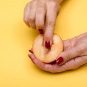Close-up of hands with red nails holding and touching a peach with a yellow background, conceptual art.