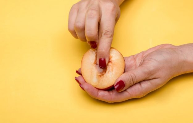 Close-up of hands with red nails holding and touching a peach with a yellow background, conceptual art.