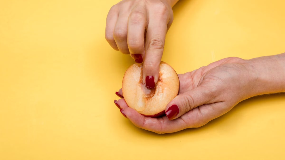 Close-up of hands with red nails holding and touching a peach with a yellow background, conceptual art.